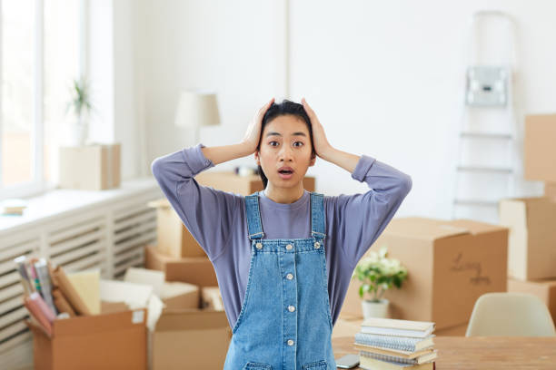 Waist up portrait of young Asian woman panicking while standing among cardboard boxes in empty room and looking at camera with big eyes, house moving or relocation concept, copy space